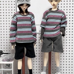 Pu&ograve; includere: Due bambole che indossano camicie a maniche lunghe a righe e pantaloncini. La bambola a sinistra indossa un berretto grigio e occhiali con montatura nera. Entrambe le bambole indossano scarpe da ginnastica bianche.