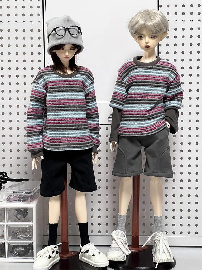 Pu&ograve; includere: Due bambole che indossano camicie a maniche lunghe a righe e pantaloncini. La bambola a sinistra indossa un berretto grigio e occhiali con montatura nera. Entrambe le bambole indossano scarpe da ginnastica bianche.