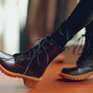 May include: A pair of brown leather boots with laces and a light brown sole. The boots have a small heel and are worn with black leggings. The boots are a classic style, suitable for various outfits.