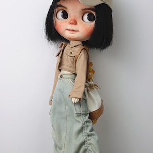 May include: A doll wearing a beige baseball cap with the word "STAR" in black, a beige cropped jacket, a light blue denim skirt, and white trainers. The doll has black hair and a backpack.