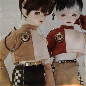 May include: Two dolls wearing crop tops with a chain detail and a letter 'B' on the front. The doll on the left is wearing a brown and white crop top with a black and white checkered skirt. The doll on the right is wearing a red and white crop top with beige trousers.