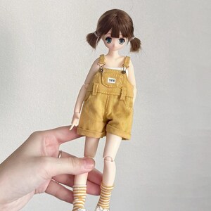 May include: A doll with brown hair styled in pigtails, wearing a yellow overall shorts set with the text "TWW" on the bib. The doll is also wearing a white tank top, striped socks, and white sneakers with black stripes.
