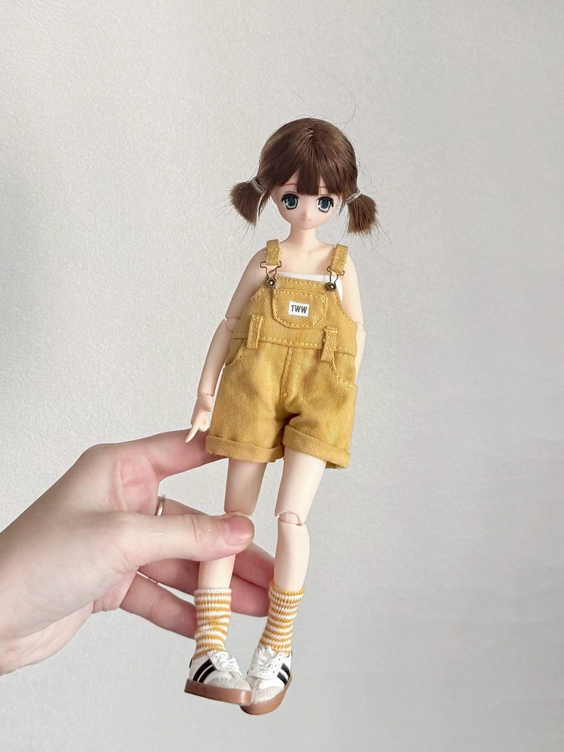 May include: A doll with brown hair styled in pigtails, wearing a yellow overall shorts set with the text "TWW" on the bib. The doll is also wearing a white tank top, striped socks, and white sneakers with black stripes.
