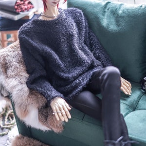 May include: A doll with red hair wearing a dark grey fuzzy sweater and black trousers. The doll is seated on a green sofa with a faux fur throw. The doll is wearing a silver necklace and rings. The doll's boots are black.