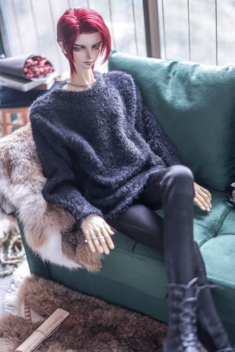 May include: A doll with red hair wearing a dark grey fuzzy sweater and black trousers. The doll is seated on a green sofa with a faux fur throw. The doll is wearing a silver necklace and rings. The doll's boots are black.