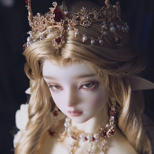 Könnte beinhalten: Eine Porzellanpuppe mit blonden Haaren, die eine goldene Krone mit roten Edelsteinen, eine goldene Halskette mit roten Edelsteinen und weißen Perlen sowie goldene Ohrringe mit roten Edelsteinen trägt.