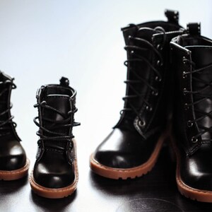 May include: Four black leather boots with brown soles are arranged in a group. Two pairs of boots are visible, one pair is smaller than the other. The boots have laces and a classic design.