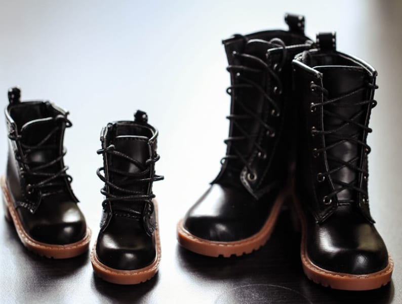 May include: Four black leather boots with brown soles are arranged in a group. Two pairs of boots are visible, one pair is smaller than the other. The boots have laces and a classic design.