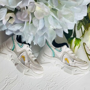 Puede incluir: Un par de zapatillas blancas con una parte superior de malla y una suela gruesa. Las zapatillas tienen un acento azul claro y amarillo.