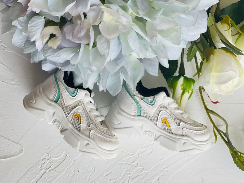 Puede incluir: Un par de zapatillas blancas con una parte superior de malla y una suela gruesa. Las zapatillas tienen un acento azul claro y amarillo.