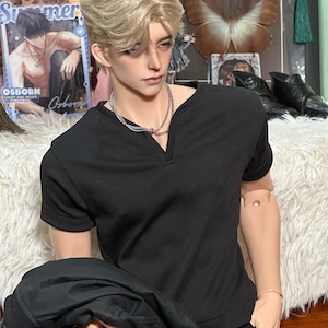 Pode incluir: Um boneco masculino com cabelo loiro usando uma camisa preta de manga curta e um colar de corrente prateada. O boneco está segurando uma jaqueta preta. No fundo, há uma foto emoldurada de um homem e uma borboleta.