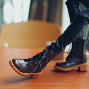 May include: A pair of brown leather boots with laces and a small heel. The boots have a light brown sole and are set against a blurred background. The boots are part of a doll's outfit.