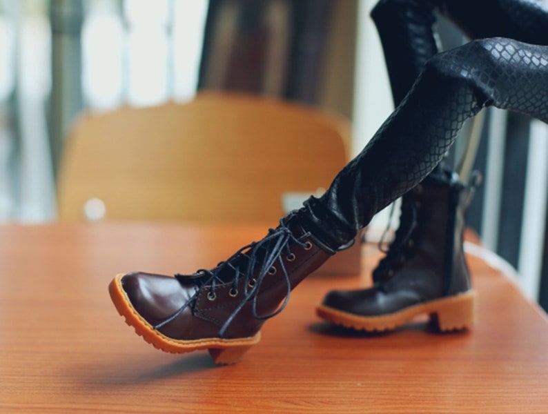 May include: A pair of brown leather boots with laces and a small heel. The boots have a light brown sole and are set against a blurred background. The boots are part of a doll's outfit.