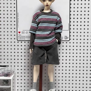 Pu&ograve; includere: Una bambola che indossa una camicia a maniche lunghe a righe con un colletto grigio scuro, pantaloncini grigi scuri e scarpe da ginnastica bianche. La bambola ha capelli corti argentati e occhi azzurri. La bambola &egrave; in piedi su un supporto di legno su uno sfondo bianco con un motivo di piccoli punti neri.