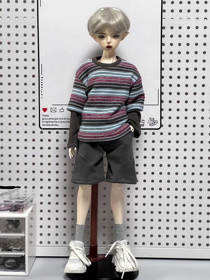 Pu&ograve; includere: Una bambola che indossa una camicia a maniche lunghe a righe con un colletto grigio scuro, pantaloncini grigi scuri e scarpe da ginnastica bianche. La bambola ha capelli corti argentati e occhi azzurri. La bambola &egrave; in piedi su un supporto di legno su uno sfondo bianco con un motivo di piccoli punti neri.