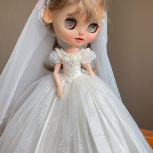 Puede incluir: Una muñeca vestida con un vestido de novia blanco con una falda amplia y adornos de encaje. La muñeca lleva un velo, un collar de perlas y una diadema con lazos rosas. La cara de la muñeca tiene ojos grandes y mejillas rosadas.