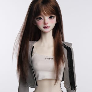 Op de afbeelding: Een pop met lang bruin haar die een grijze rits-hoodie draagt met de tekst "SWEET 77" op een wit tanktop eronder.