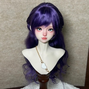 May include: A ball-jointed doll head and shoulders bust featuring long, wavy purple hair with bangs. The doll has pale skin, large dark eyes, and a soft pink blush. A gold-toned chain necklace with a single white heart-shaped pendant is worn.