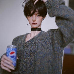 May include: A doll wearing a blue and brown knitted jumper with a black choker necklace and a silver earring. The doll is holding a can of Pepsi.