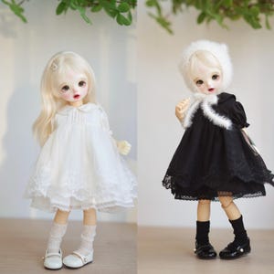 May include: Two dolls wearing different outfits. The doll on the left is wearing a white dress with lace trim and white socks. The doll on the right is wearing a black dress with lace trim and black socks. Both dolls have blonde hair and are wearing white shoes.