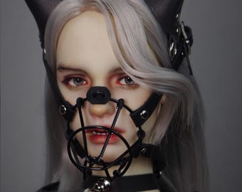 May include: A doll wearing a black leather muzzle with a metal cage and a black leather choker with a silver chain attached. The doll has long, silver hair and is wearing a black leather jacket.