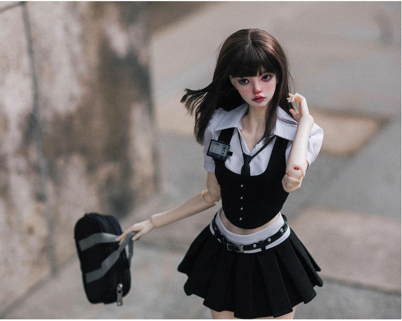 May include: A doll dressed in a black and white school uniform, including a pleated skirt, vest, and collared shirt. The doll has dark hair and is holding a black backpack. A small ID badge is pinned to the shirt.