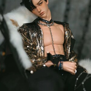 Op de afbeelding: Een pop met kort zwart haar die een gouden paillettenjas, een zwarte broek en een zilveren kettingketting draagt.