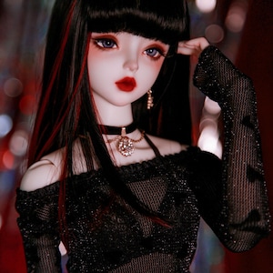 May include: A doll with long black hair with red streaks, wearing a black mesh top with a black choker and a gold chain necklace with a large rhinestone pendant. The doll has a white face with red lips and blue eyes.