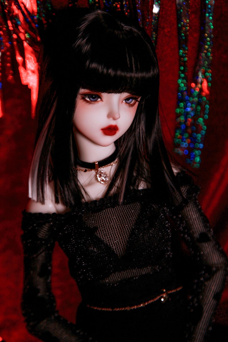 May include: A doll with long black hair and bangs, wearing a black off-the-shoulder top with a mesh overlay and a gold belt. The doll has blue eyes and red lips. The background is a red fabric with a blurry pattern of colorful lights.