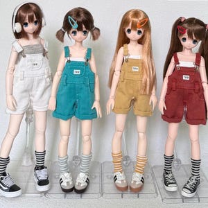 May include: Four fashion dolls wearing different colored overalls and sneakers. The dolls have various hairstyles and accessories, including headphones and hair clips. Each doll is standing on a clear plastic stand.