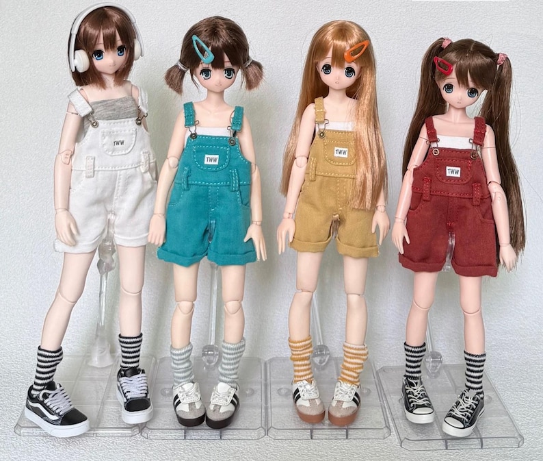 May include: Four fashion dolls wearing different colored overalls and sneakers. The dolls have various hairstyles and accessories, including headphones and hair clips. Each doll is standing on a clear plastic stand.