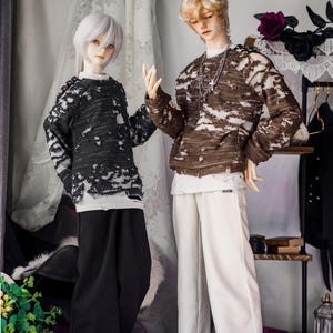 May include: Two fashion dolls are posed, showcasing distressed-style sweaters and wide-leg trousers. One doll wears a black sweater over a white shirt and black trousers. The other doll is in a brown sweater, white shirt, and white trousers. Both dolls have light-coloured hair.