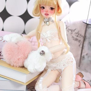 May include: A doll with blonde hair and cat ears wearing a white lace lingerie set with a pink fluffy toy. The doll is sitting on a white surface with a book and a piece of paper with writing on it.
