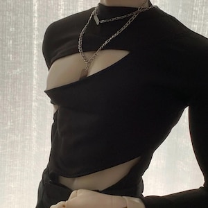 May include: Black long-sleeved top with a unique cut-out design. The top features a high neckline and long sleeves. A silver chain necklace with a rectangular pendant is worn. The top is made of a soft, stretchy material.