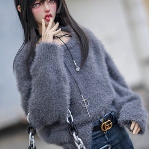 May include: A doll in a grey fluffy jumper, dark blue jeans, and a black belt with a gold buckle. The doll has long black hair and is holding a grey fluffy purse with a silver chain. A black necklace with a silver pendant is also visible.