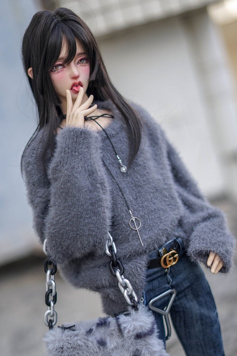 May include: A doll in a grey fluffy jumper, dark blue jeans, and a black belt with a gold buckle. The doll has long black hair and is holding a grey fluffy purse with a silver chain. A black necklace with a silver pendant is also visible.