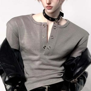 Può includere: Una bambola snodabile con capelli corti rosa chiaro, che indossa una maglietta henley grigia a maniche corte con bottoni argentati, una giacca in similpelle nera sulle spalle, un girocollo con borchie e pantaloni neri con fibbia argentata.