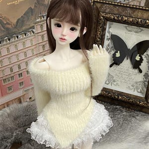 Puede incluir: Una mu&ntilde;eca de peluche blanco con un su&eacute;ter blanco peludo con un borde de encaje blanco. La mu&ntilde;eca tiene el pelo largo y casta&ntilde;o y lleva una falda de encaje blanco.