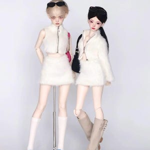 May include: Two fashion dolls stand against a white background. They wear matching white fluffy crop tops and skirts. One doll has blonde hair and blue trainers, the other has black hair and tan boots. Both wear sunglasses and carry handbags.