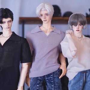 May include: Three dolls wearing different colored shirts and jeans. The doll on the left is wearing a black shirt, the doll in the middle is wearing a lavender shirt, and the doll on the right is wearing a white shirt.