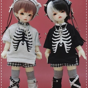 May include: Two dolls wearing black and white striped socks and checked shorts. One doll is wearing a grey shirt with a skeleton print and a grey and white hat. The other doll is wearing a black shirt with a skeleton print and a black and white hat.