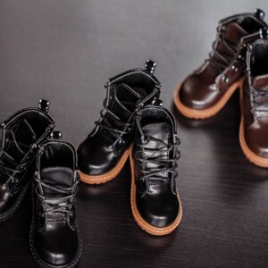 May include: Several pairs of miniature boots in black and brown leather are arranged on a dark surface. The boots have laces, rounded toes, and tan soles. The leather has a glossy finish.