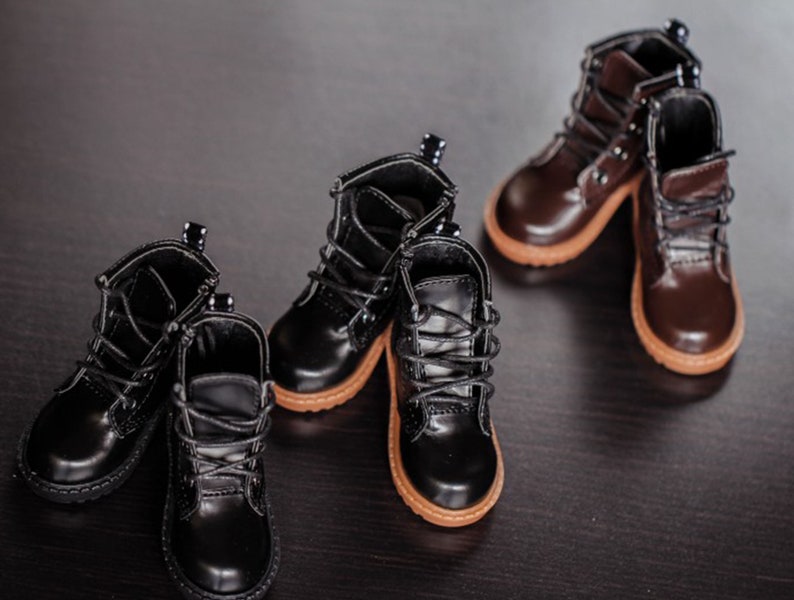 May include: Several pairs of miniature boots in black and brown leather are arranged on a dark surface. The boots have laces, rounded toes, and tan soles. The leather has a glossy finish.