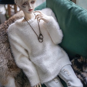 May include: A doll with pale skin, platinum blonde hair, and a white fuzzy sweater. The doll wears a dark metal handcuff pendant necklace and light-wash ripped jeans. The doll is seated on a green cushion and a fur throw.