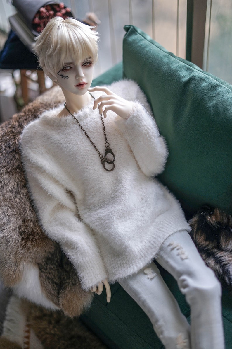 May include: A doll with pale skin, platinum blonde hair, and a white fuzzy sweater. The doll wears a dark metal handcuff pendant necklace and light-wash ripped jeans. The doll is seated on a green cushion and a fur throw.