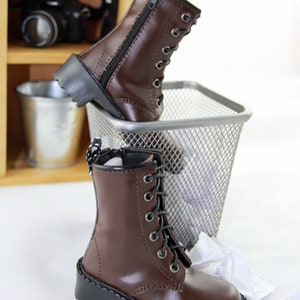 May include: Brown leather boots with black laces and zippers. The boots have a classic combat boot design with a rounded toe and a chunky black sole. The boots are displayed near a metal trash bin and a wooden shelf.