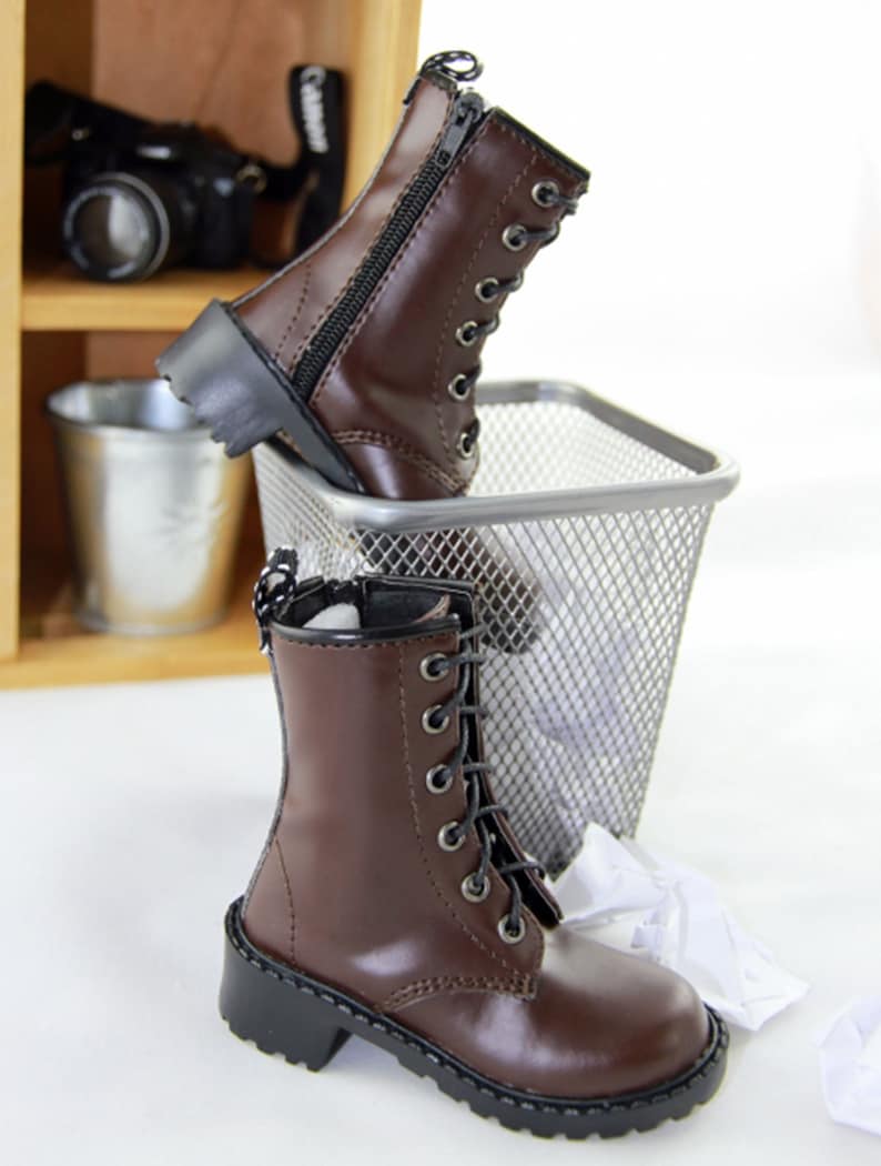 May include: Brown leather boots with black laces and zippers. The boots have a classic combat boot design with a rounded toe and a chunky black sole. The boots are displayed near a metal trash bin and a wooden shelf.