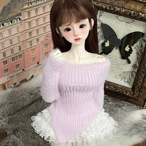 Puede incluir: Una mu&ntilde;eca con un su&eacute;ter de color lila suave y esponjoso con una falda de encaje blanco con volantes.