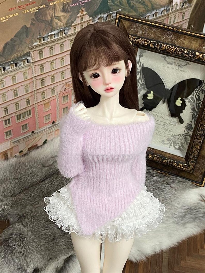Puede incluir: Una mu&ntilde;eca con un su&eacute;ter de color lila suave y esponjoso con una falda de encaje blanco con volantes.