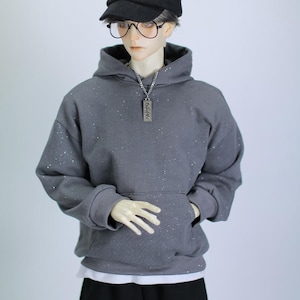 May include: A gray hoodie with a white star on the hood and a silver chain necklace with a silver tag. The hoodie has a small white dot pattern. The model is wearing a black baseball cap with a white "NY" logo and black pants.